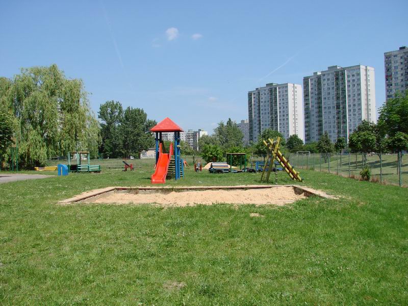 Plac zabaw, zielona trawa, duża piaskowmica, czerwona zjeżdżalnia z daszkiem, drabink,a w oddali bloki mieszkalne