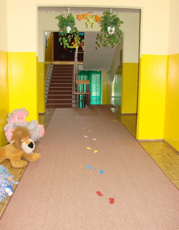 korytarz z wykładziną, po lewej stronie duże pluszaki, nad korytarzem wiszą kwiaty, z tyłu schody na piętro i schody do wyjścia