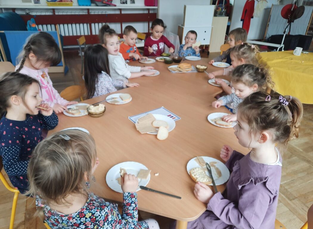 Dzieci siedzą przy stole i jedzą kanapki, na środku stołu lezy tależyk z chlebem