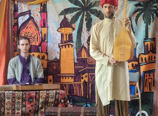 mężczyzna w stroju ludowym pokazuje turecki instrument ludowy, za nim drugi mężczyzna siedzi na tle kolorowego materiału