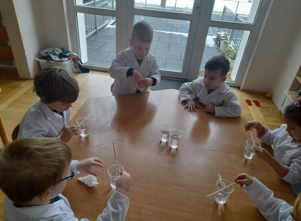 dzieci ubrane w białe fartuchy siedzą przy stole ze szklankami z wodą