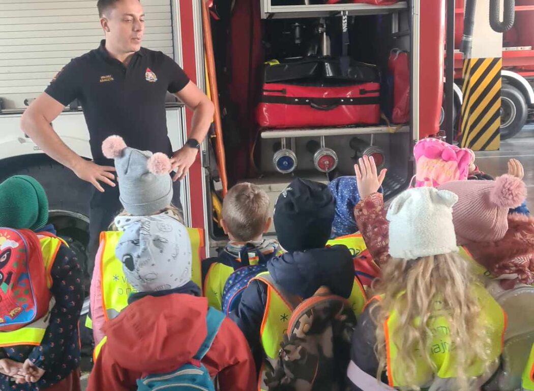 dzieci przygladają sie wozowi strażackiemu, obok stoi strażak podpierając się rękoma