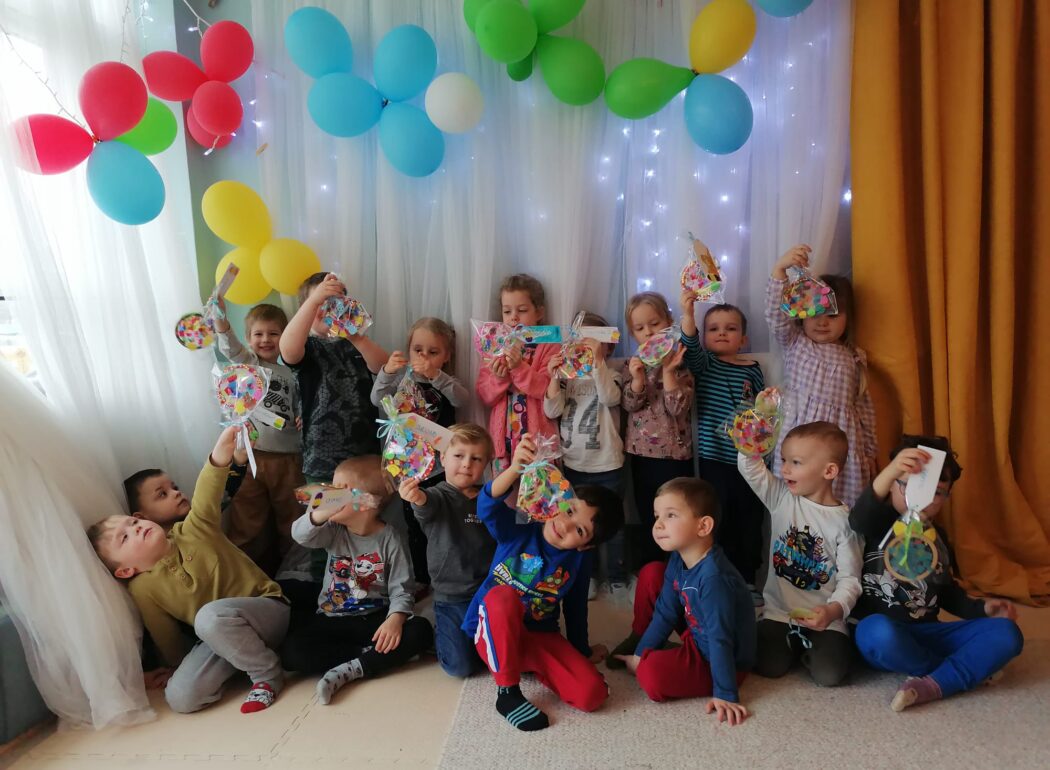 uśmiechnięte dzieci na tle ściany udekorowanej kolorowymi balonami trzymają w rękach kolorowe woreczki